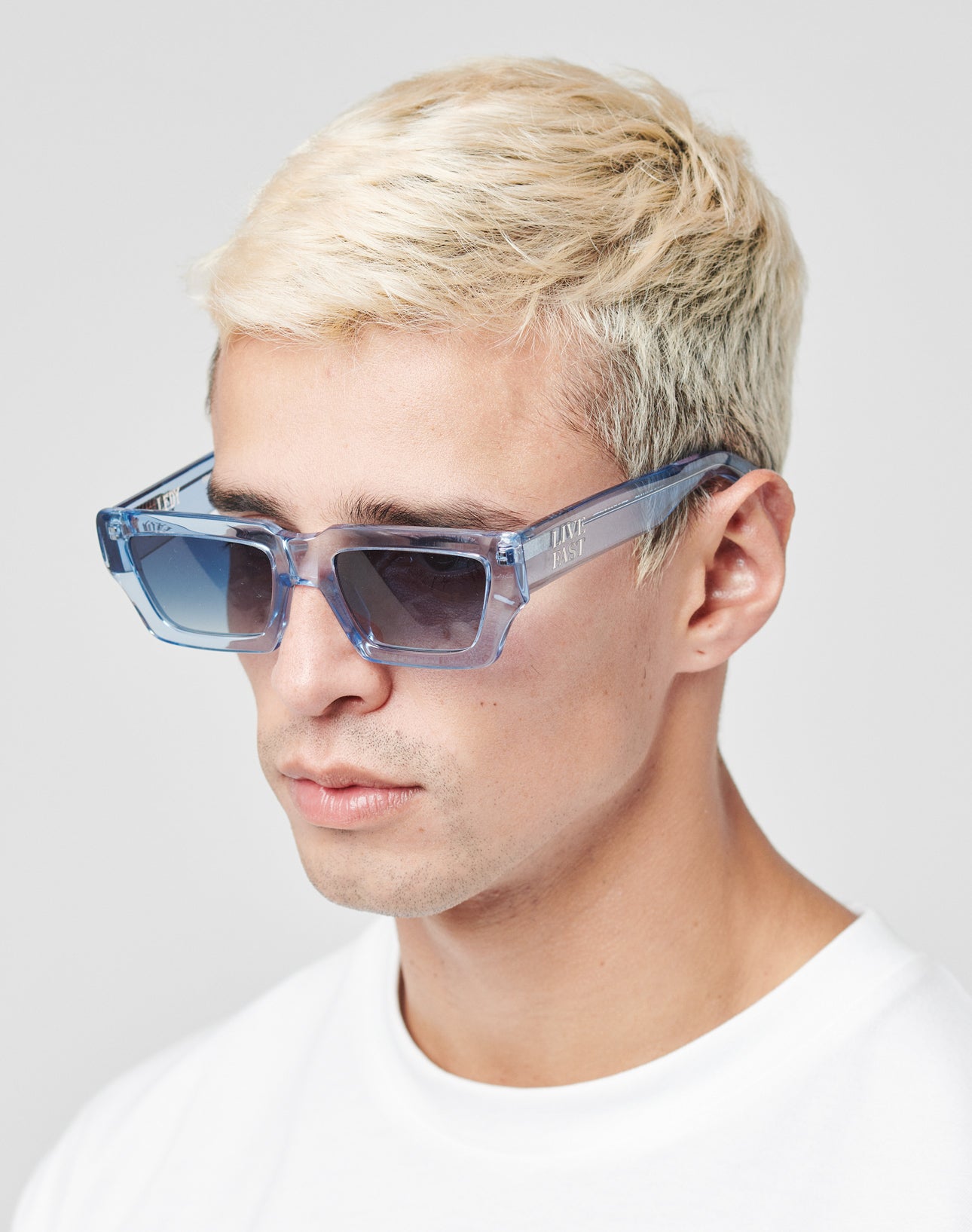 A young man with short, bleached blonde hair wears LIVE FAST DIE YOUNGs Live Fast Sunglasses—angular blue frames crafted from premium Mazzuchelli Acetate—paired with a white shirt, gazing downward against a plain light background.
