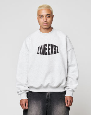 A person with short blond hair wears the LFDY Emblem Sweater by LIVE FAST DIE YOUNG, an oversized light gray cotton sweatshirt with bold black LIVE FAST lettering, paired with dark jeans against a plain light background.
