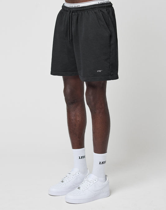 A person wears LIVE FAST DIE YOUNG Basic Sweat Shorts with an elastic waistband and LFTV logo print, paired with white socks and a waistband featuring the LFTV text, showcasing a relaxed, oversized fit against a plain white background.