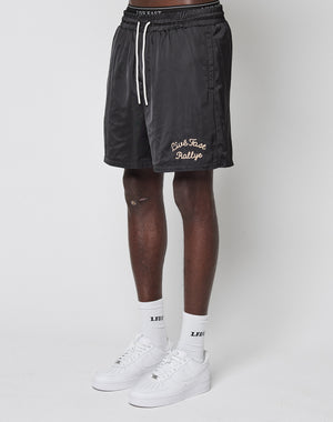 A person wearing LIVE FAST DIE YOUNG’s LFDY Retro Satin Shorts in black with a beige chainstitch logo, white LFDY socks, and white sneakers stands against a plain white background.
