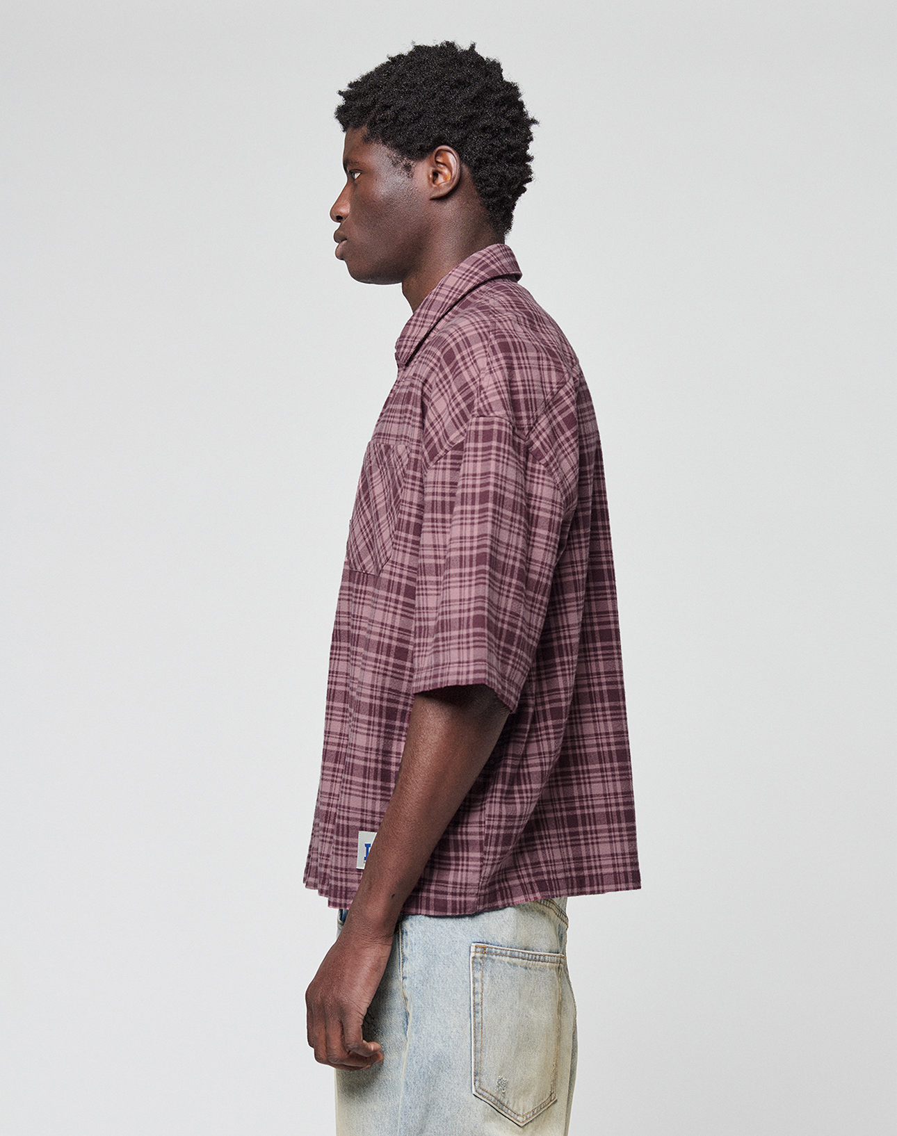 A young man stands in profile against a plain background, wearing the LFDY Light Flannel in maroon plaid from LIVE FAST DIE YOUNG and light-wash jeans, both crafted from 100% cotton for comfort and style.