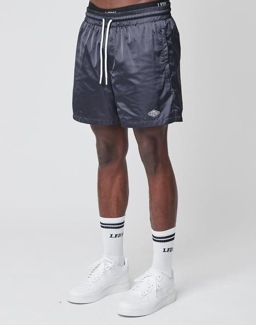 A person in LIVE FAST DIE YOUNG Basic Satin Shorts stands against a plain light background, paired with white crew socks featuring black stripes and white sneakers.