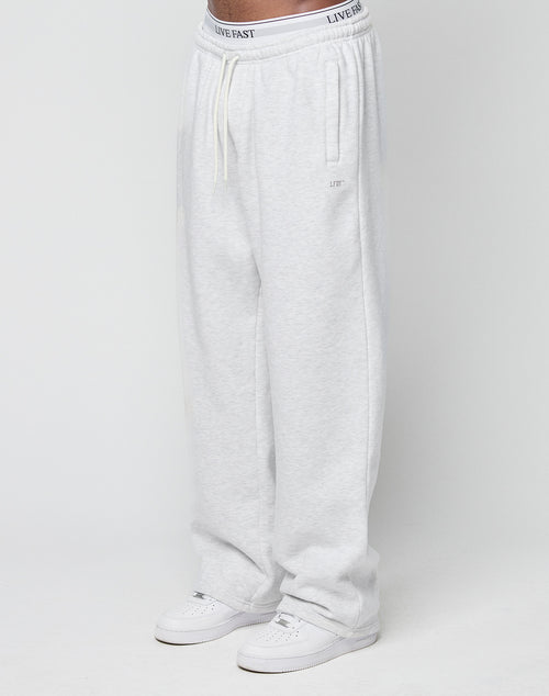 A person wearing Basic Wide Jogger sweatpants by LIVE FAST DIE YOUNG in light gray with an elastic waistband, drawstring, and bold logo print, paired with white sneakers, stands against a plain white background.