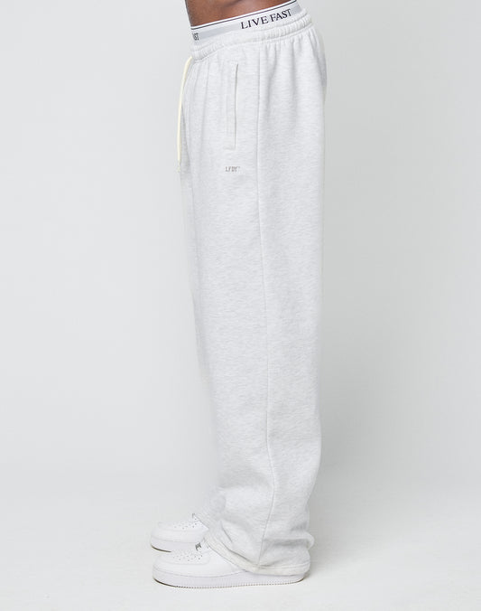 A person in profile wears LIVE FAST DIE YOUNG Basic Wide Jogger sweatpants in light gray, featuring a relaxed fit, subtle upper thigh logo, and elastic waistband, styled with white sneakers against a plain white background.