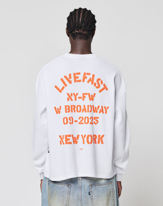 Seen from the back, a person with braided hair wears the LIVE FAST DIE YOUNG NY-FW Waffle Longsleeve—a white, 100% Baumwolle oversized shirt with bold orange “LIVE FAST NYC-FW W BROADWAY 09-2025 NEW YORK” backprint—paired with faded blue jeans.