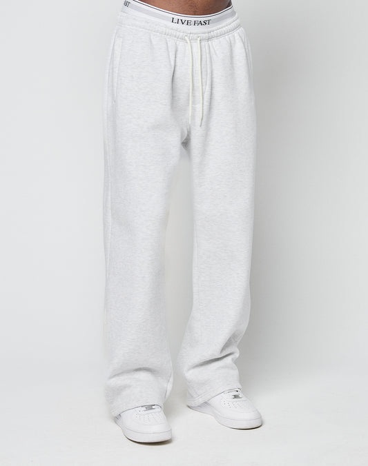 A person wearing LIVE FAST DIE YOUNG Basic Wide Jogger sweatpants in light gray with an elastic waistband and drawstring, white sneakers, and underwear with LIVE FAST printed on the elastic band, standing before a plain white background.