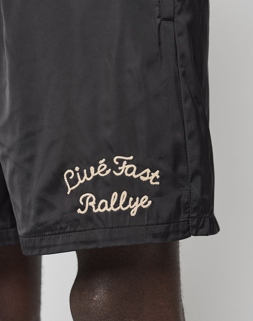 A close-up of someone wearing LFDY Retro Satin Shorts by LIVE FAST DIE YOUNG, featuring an oversize fit and beige chainstitch Live Fast Rallye logo on the lower left leg.