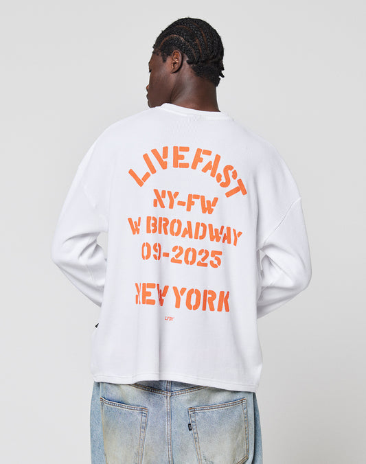 Wearing the LIVE FAST DIE YOUNG NY-FW Waffle Longsleeve, an oversize white 100% cotton shirt with a bold orange backprint: LIVE FAST XY-FW W BROADWAY 09-2025 NEW YORK. The short-haired model faces away from the camera.