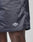 Close-up of a person’s dark-skinned leg wearing LIVE FAST DIE YOUNG Basic Satin Shorts in dark gray with a soft polyester feel and a small logo patch near the hem, set against a plain, light background.