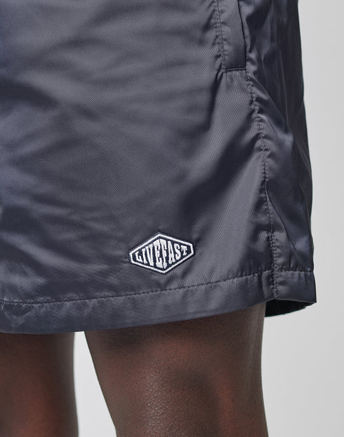 Close-up of a person’s dark-skinned leg wearing LIVE FAST DIE YOUNG Basic Satin Shorts in dark gray with a soft polyester feel and a small logo patch near the hem, set against a plain, light background.