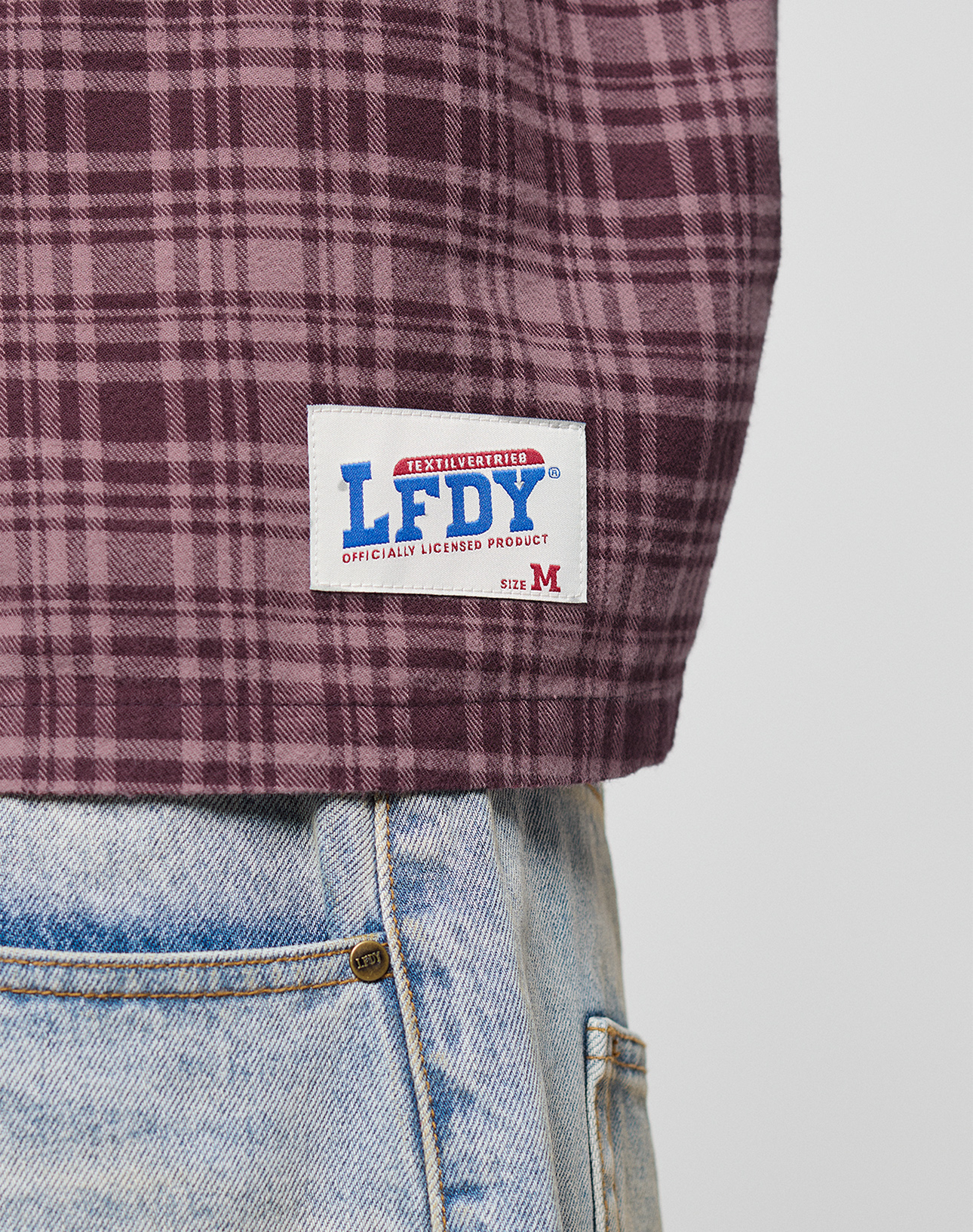 A close-up of a person in blue jeans and an LFDY Light Flannel by LIVE FAST DIE YOUNG in maroon plaid, with LFDY branding and a white LFDY Size M patch near the hem. Made from 100% Baumwolle for comfort.