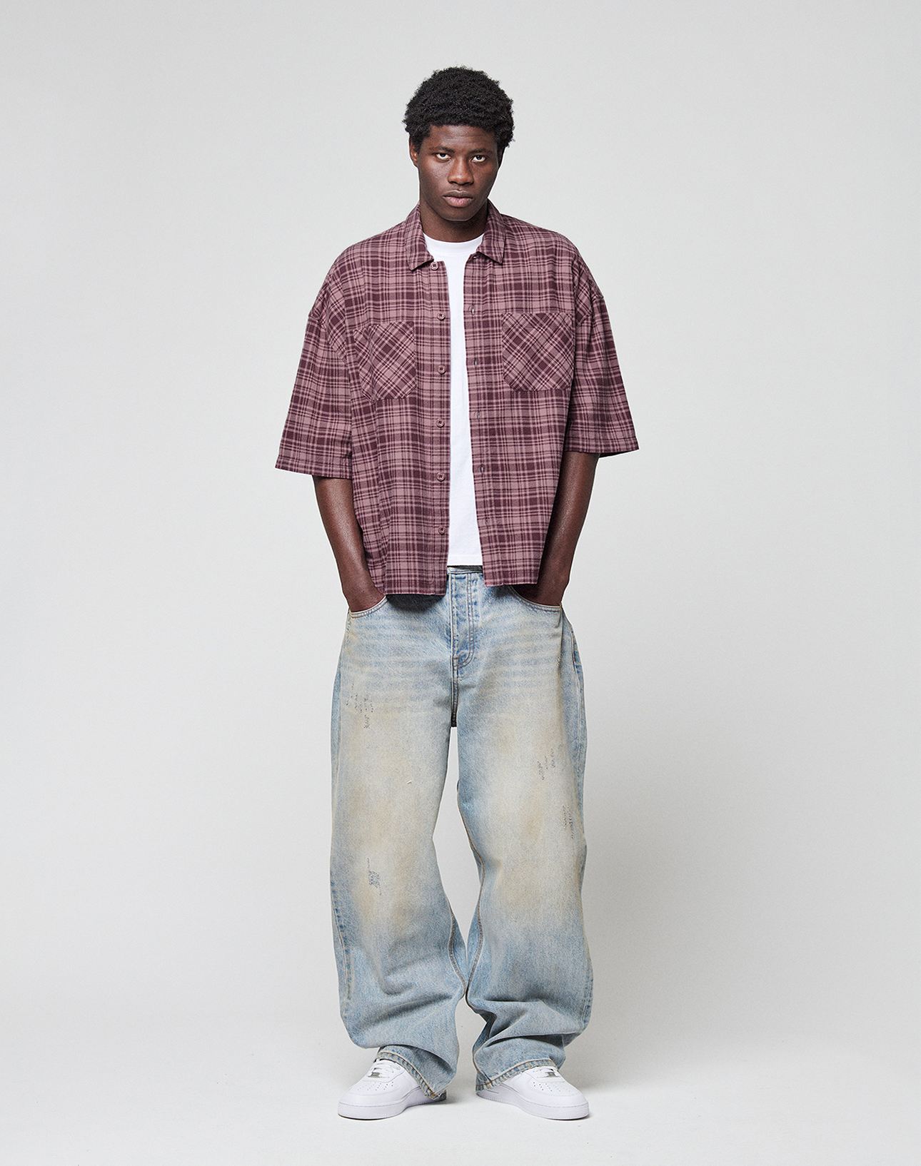 A man stands against a plain background, wearing the maroon plaid LFDY Light Flannel by LIVE FAST DIE YOUNG over a white tee. He pairs it with loose faded blue jeans and white sneakers. The shirt is crafted from 100% cotton for comfort.