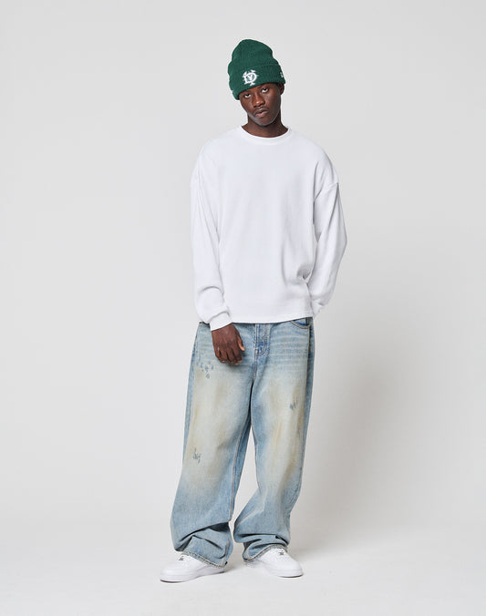 A man in a green beanie, LIVE FAST DIE YOUNG NY-FW Waffle Longsleeve (white, oversize fit, 100% cotton) with backprint, baggy light-wash jeans, and white sneakers stands relaxed against a plain white background.