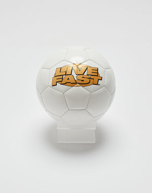 The Live Fast Soccer Ball by LIVE FAST DIE YOUNG features black stitching, bold yellow and black LIVE FAST lettering, and a distinctive logo print, all displayed on a clear stand against a plain white background.