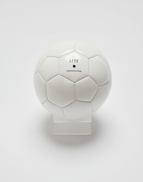 A white Live Fast Soccer Ball by LIVE FAST DIE YOUNG with black stitched panels sits on a transparent stand, featuring LFDY, INDOOR AND OUTDOOR, and a Logo Aufdruck in black on its plain surface.