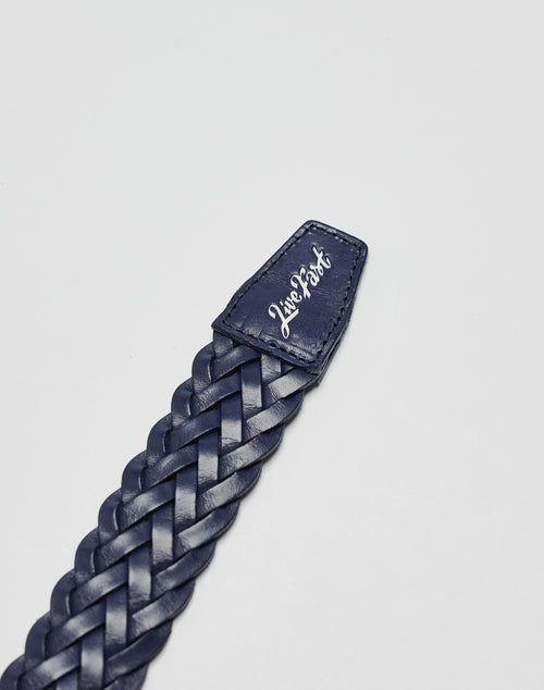A close-up of the navy blue LFDY Braided Belt by LIVE FAST DIE YOUNG, made from 100% cow leather, with a stitched end reading “Live Fast” in white cursive and a silver buckle, set on a plain light gray background.