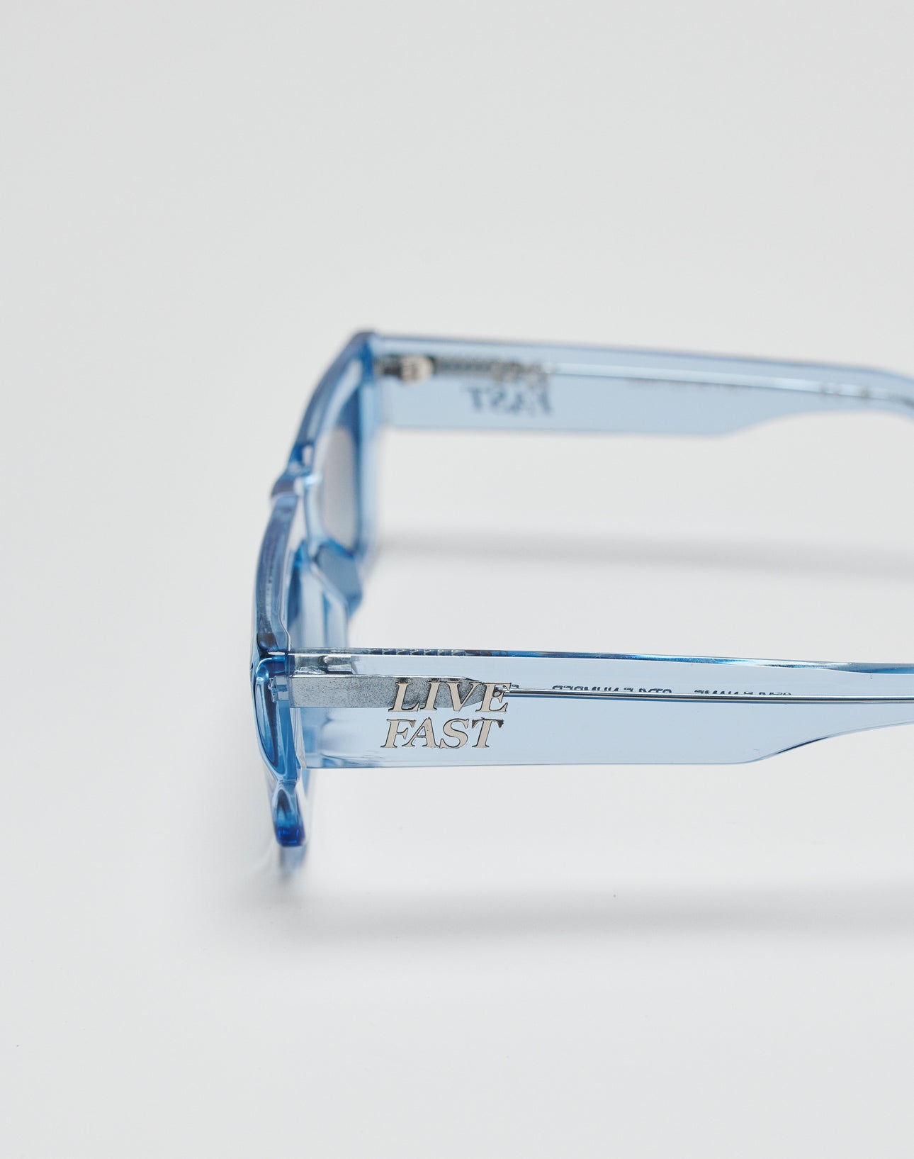 Close-up of transparent blue Live Fast Sunglasses by LIVE FAST DIE YOUNG, featuring LIVE FAST engraved in silver on the side, shown against a plain white background.
