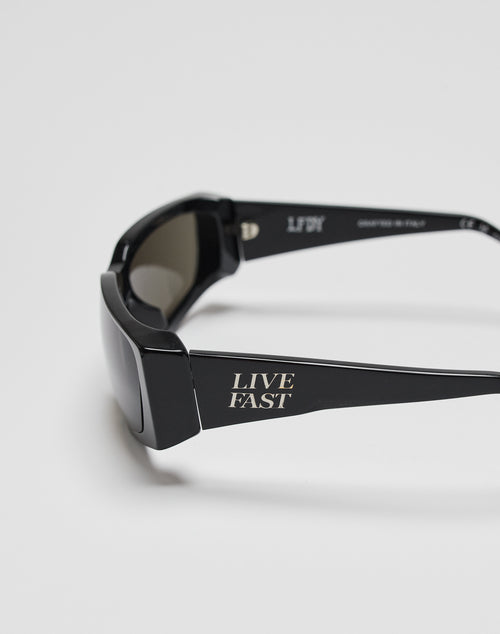 Live Fast Curved Shades by LIVE FAST DIE YOUNG: Black sunglasses with thick arms, LIVE FAST printed in white, crafted from Mazzuchelli Acetat with 400 UV protection. Made in Italy. Shown on a plain, light-colored background.