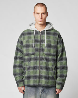 A person with short hair wears a green and black LIVE FAST DIE YOUNG Vintage Padded Hooded Flannel over a gray sweatshirt and light blue vintage wash jeans, standing against a plain light background.