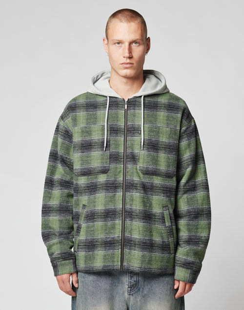 A person with short hair wears a green and black LIVE FAST DIE YOUNG Vintage Padded Hooded Flannel over a gray sweatshirt and light blue vintage wash jeans, standing against a plain light background.