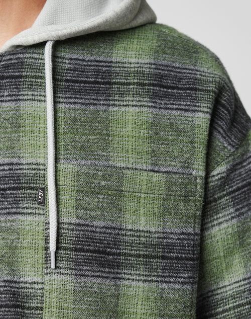Close-up of the LIVE FAST DIE YOUNG Vintage Padded Hooded Flannel in green and black plaid with a light gray hood, drawstring, and visible Flag-Label tag, showing part of a person’s shoulder and chest.