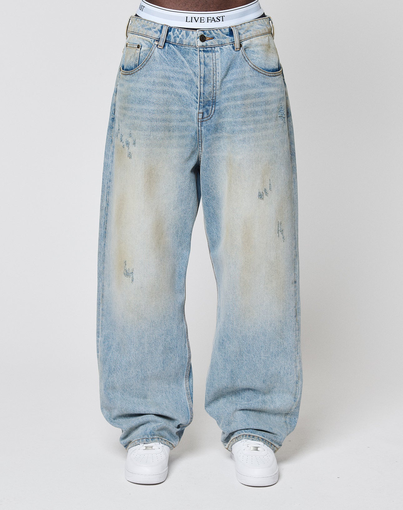 A person wears LFDY Baggy Denim by LIVE FAST DIE YOUNG—light-wash, distressed, with slight fading—paired with white sneakers and visible LIVE FAST waistband. The background is plain gray.