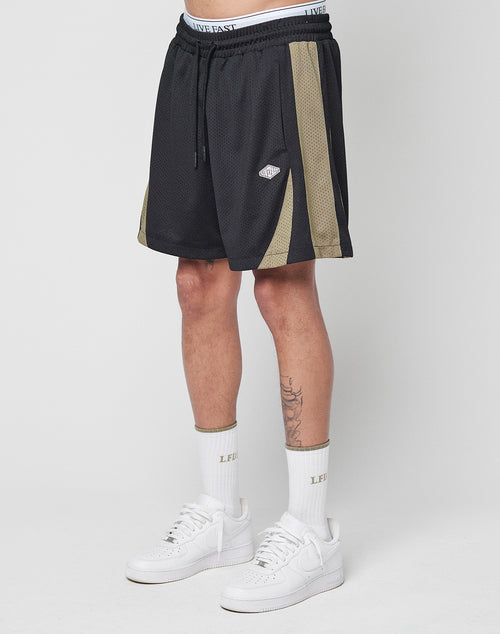 A person stands against a white background wearing LIVE FAST DIE YOUNG Striped Mesh Shorts—black with gold side stripes, white logo patch—paired with LIVE FAST text crew socks and white sneakers. The shorts have a regular fit and comfortable polyester feel.