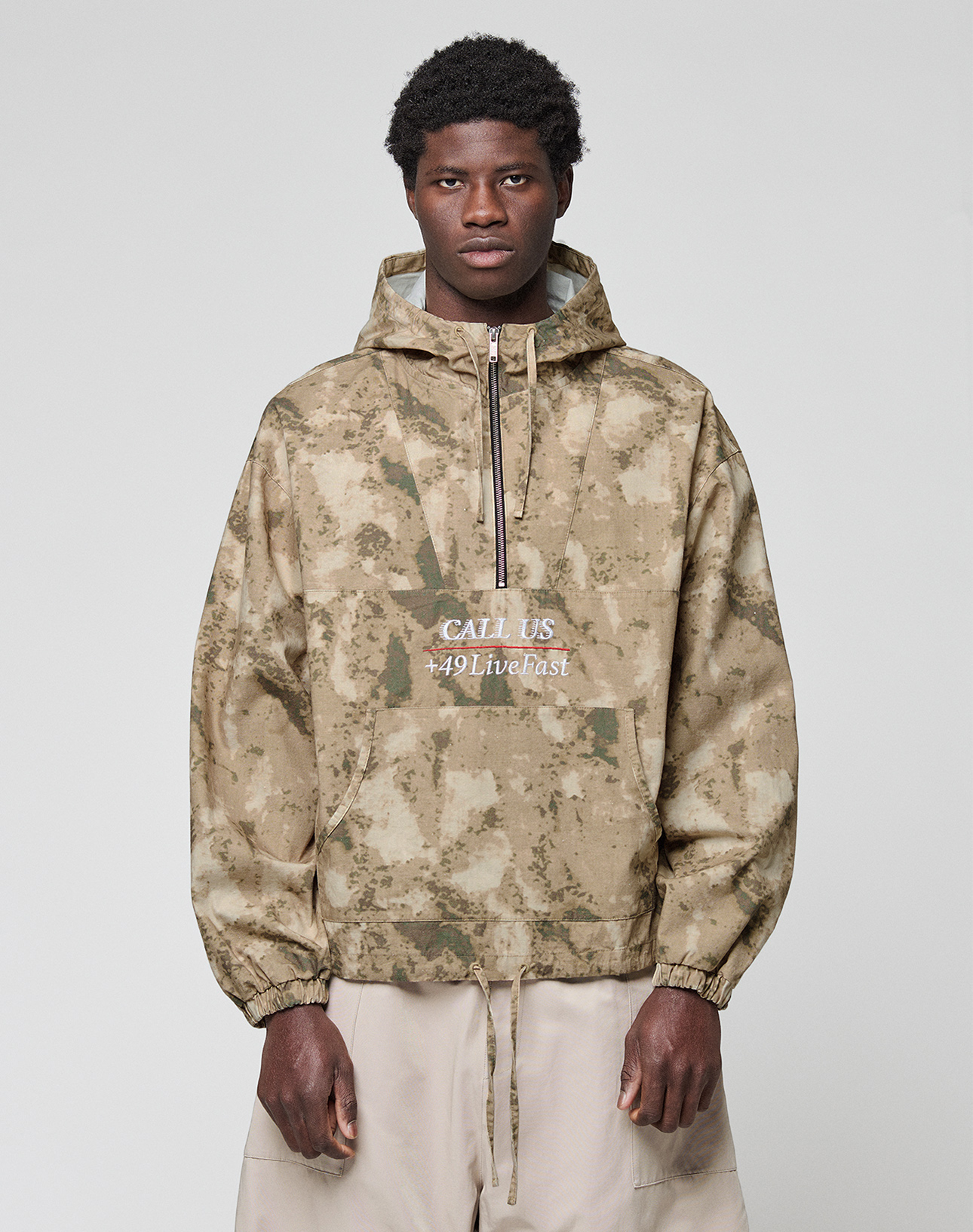 A man with short curly hair wears a LIVE FAST DIE YOUNG LFDY Camo Halfzip Jacket in oversize fit with "CALL US 449Livefast" on the front, paired with light beige 100% Baumwolle pants, standing against a light gray backdrop.