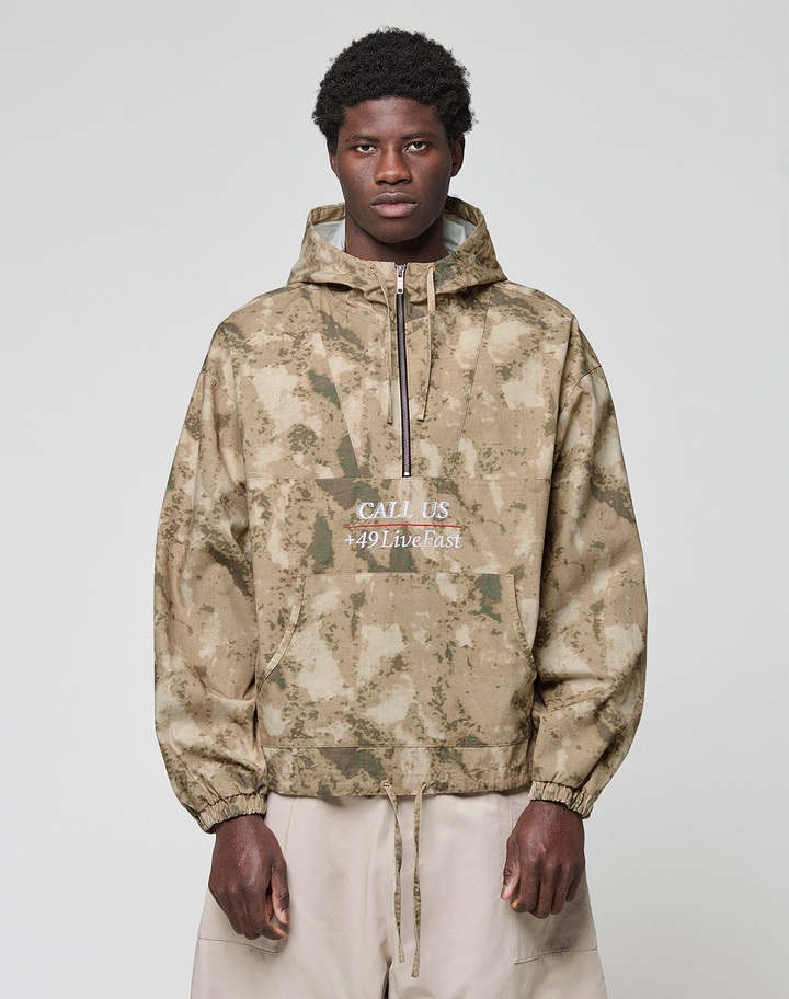 A man with short curly hair wears a LIVE FAST DIE YOUNG LFDY Camo Halfzip Jacket in oversize fit with "CALL US 449Livefast" on the front, paired with light beige 100% Baumwolle pants, standing against a light gray backdrop.
