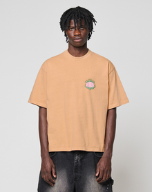 A man with medium-length twisted hair wears the LIVE FAST DIE YOUNG LFDY Flames Chest Tee, a regular fit peach shirt with small front embroidery, and faded black jeans. He stands against a plain light gray background, facing the camera.