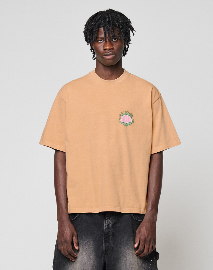 A man with medium-length twisted hair wears the LIVE FAST DIE YOUNG LFDY Flames Chest Tee, a regular fit peach shirt with small front embroidery, and faded black jeans. He stands against a plain light gray background, facing the camera.