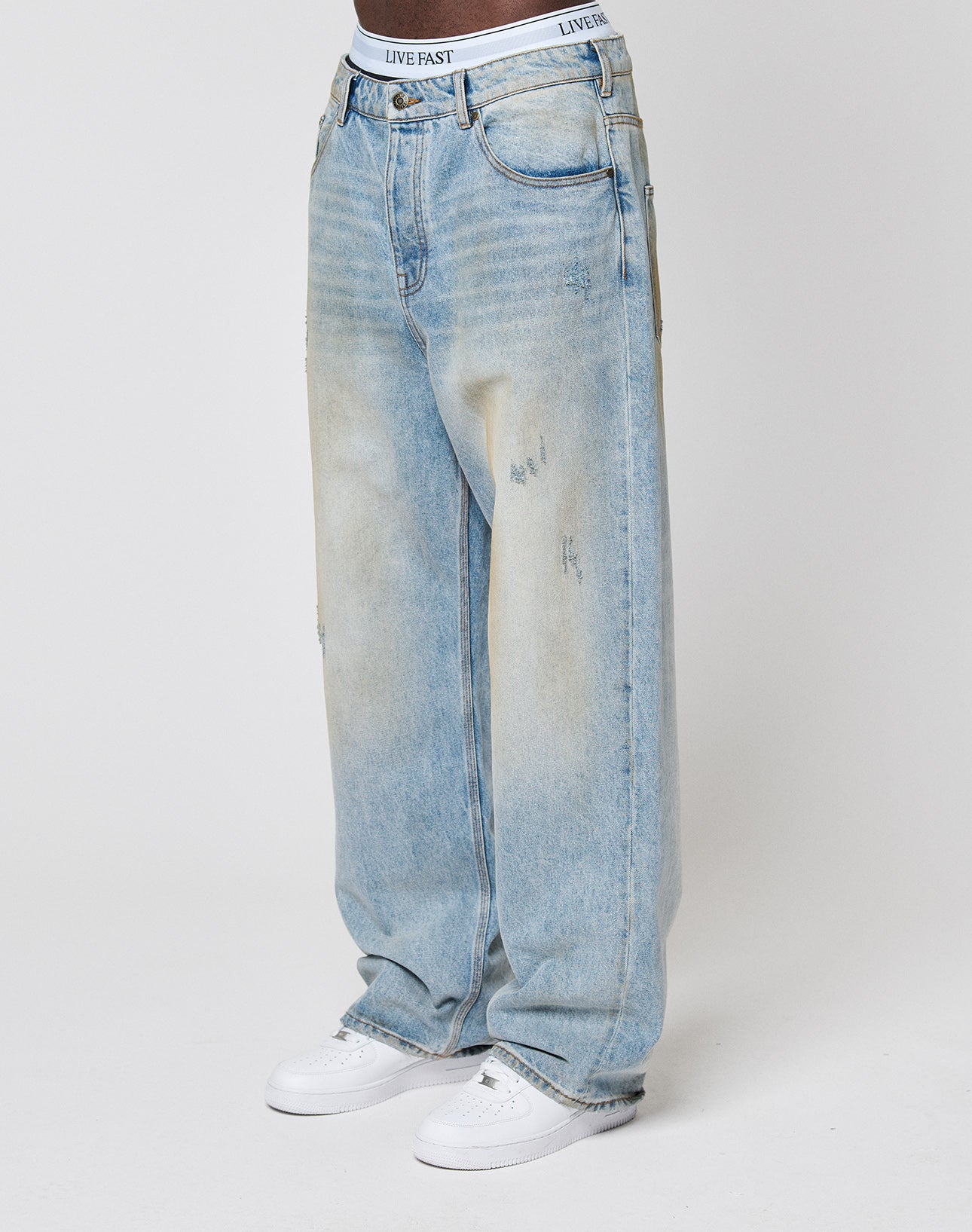 A person wears LFDY Baggy Denim by LIVE FAST DIE YOUNG—light-wash, subtly distressed jeans—with white sneakers and a white waistband featuring LIVE FAST branding, standing against a plain white background.