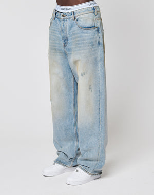 A person wears LFDY Baggy Denim by LIVE FAST DIE YOUNG—light-wash, subtly distressed jeans—with white sneakers and a white waistband featuring LIVE FAST branding, standing against a plain white background.