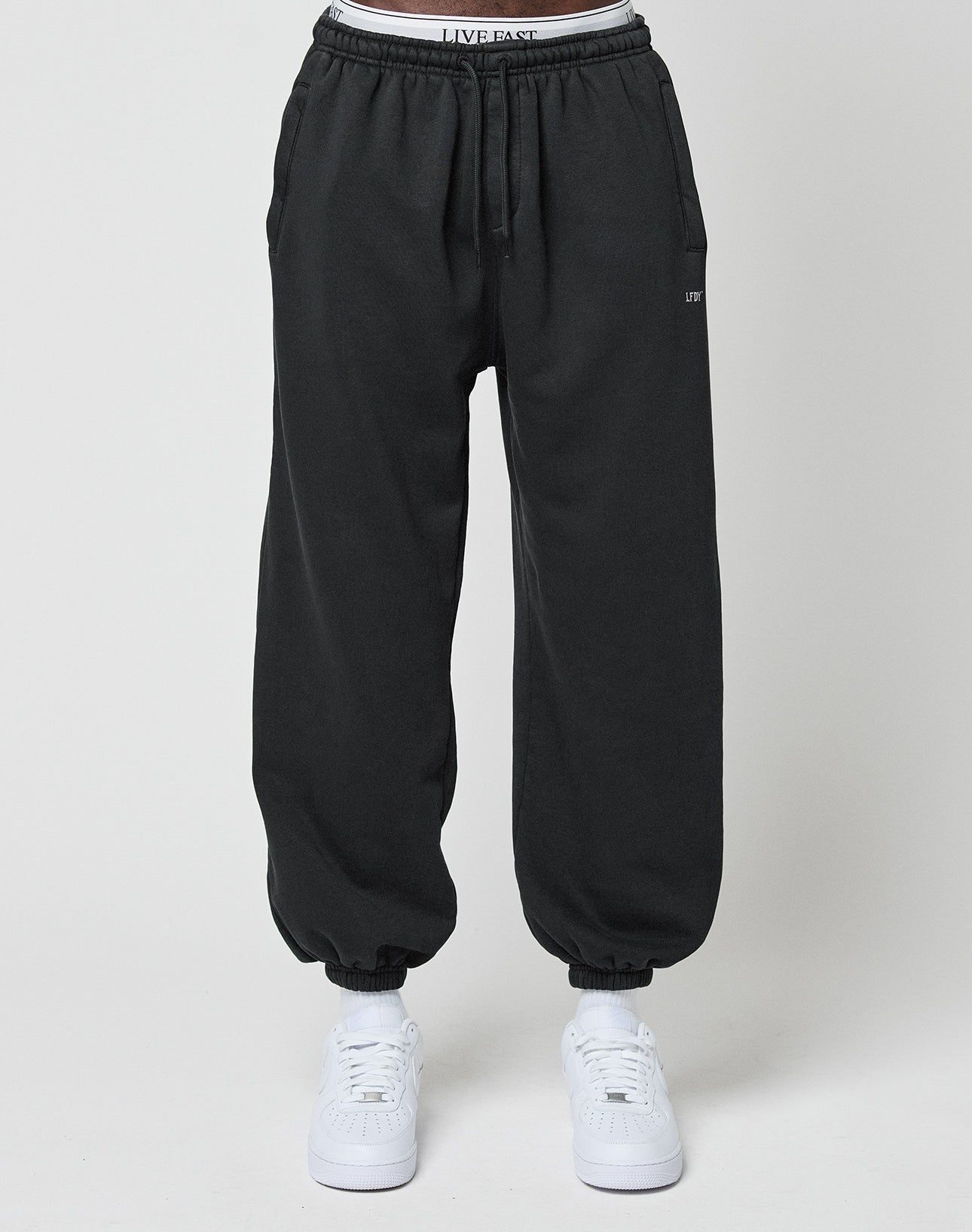 Someone wears the LIVE FAST DIE YOUNG Basic Loose Jogger in black, featuring an elastic waistband and cuffs, with LIVE FAST visible on the waist. They pair it with white sneakers and stand before a plain light background.