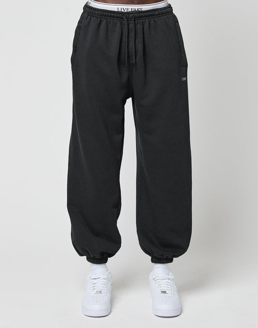 Someone wears the LIVE FAST DIE YOUNG Basic Loose Jogger in black, featuring an elastic waistband and cuffs, with LIVE FAST visible on the waist. They pair it with white sneakers and stand before a plain light background.