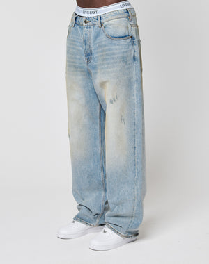 A person wears light blue LFDY Baggy Dirt Denim jeans by LIVE FAST DIE YOUNG, featuring a relaxed, baggy fit, subtle front fading, gerades Bein, and a visible white LIVE FAST waistband, paired with white sneakers. Background is plain white.