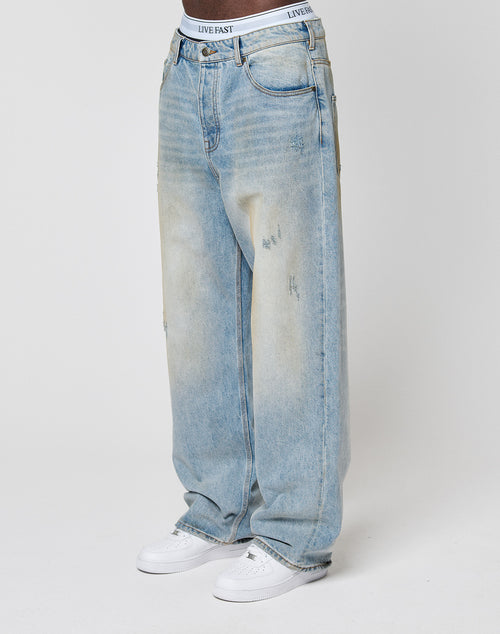 A person wears light blue LFDY Baggy Dirt Denim jeans by LIVE FAST DIE YOUNG, featuring a relaxed, baggy fit, subtle front fading, gerades Bein, and a visible white LIVE FAST waistband, paired with white sneakers. Background is plain white.