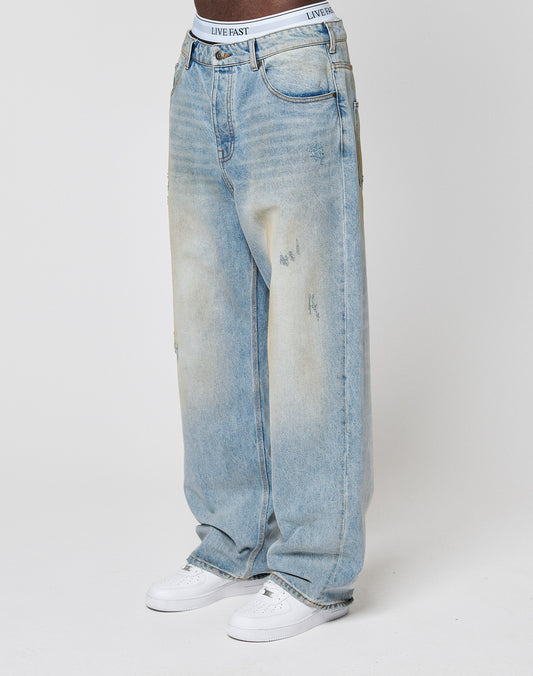 A person wears Baggy Dirt Denim jeans from LIVE FAST DIE YOUNG—loose-fitting, light blue, distressed, with a white LIVE FAST waistband—paired with white sneakers, standing against a plain light background.