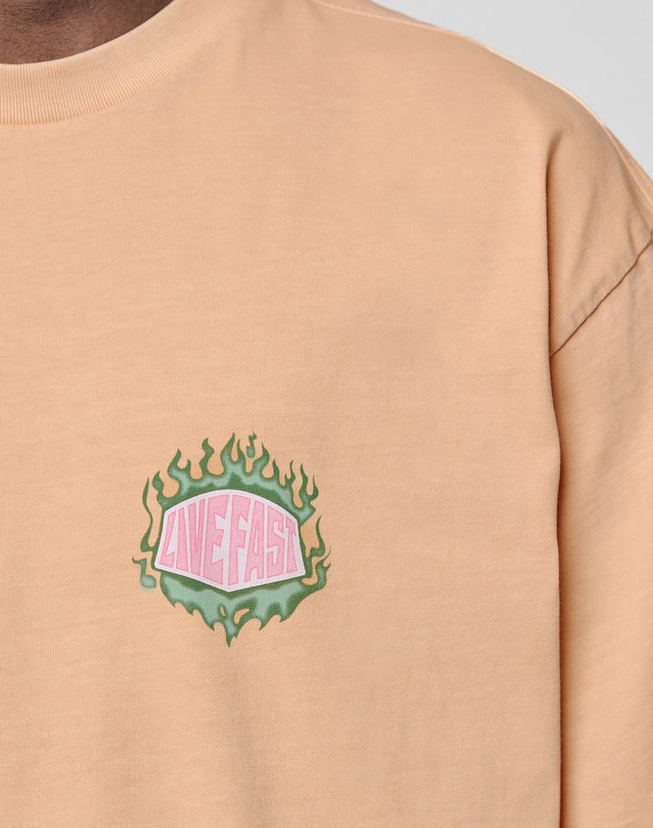 A close-up of the LFDY Flames Chest Tee by LIVE FAST DIE YOUNG, a regular fit peach shirt in 100% cotton, features a small green flame front print with "LOVE FAST" in pink and white on the left chest.