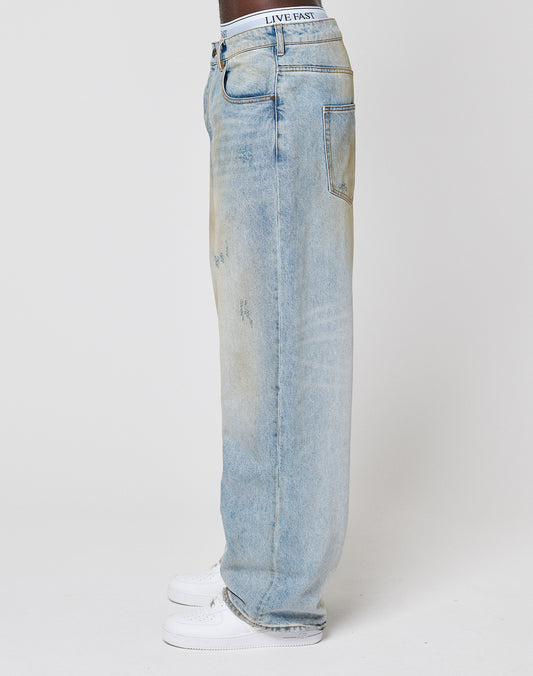 A person stands sideways in LFDY Baggy Denim by LIVE FAST DIE YOUNG—light-wash blue jeans with subtle distressing and a LIVE FAST waistband—paired with white sneakers against a plain white background.