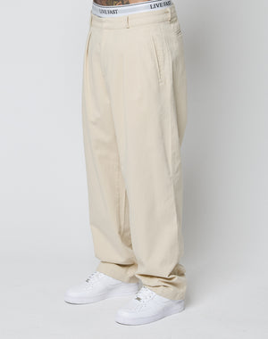 Model in beige Wide Chino trousers (100% Baumwolle, Doppel-Bundfalte) by LIVE FAST DIE YOUNG, with visible white LIVE FAST waistband underwear and white sneakers, standing against a plain white background.