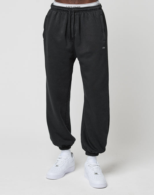 A person wears LIVE FAST DIE YOUNG Basic Loose Jogger sweatpants with Elastic-Bund cuffs and a loose fit, paired with white sneakers and a visible white waistband, standing against a plain light gray background.