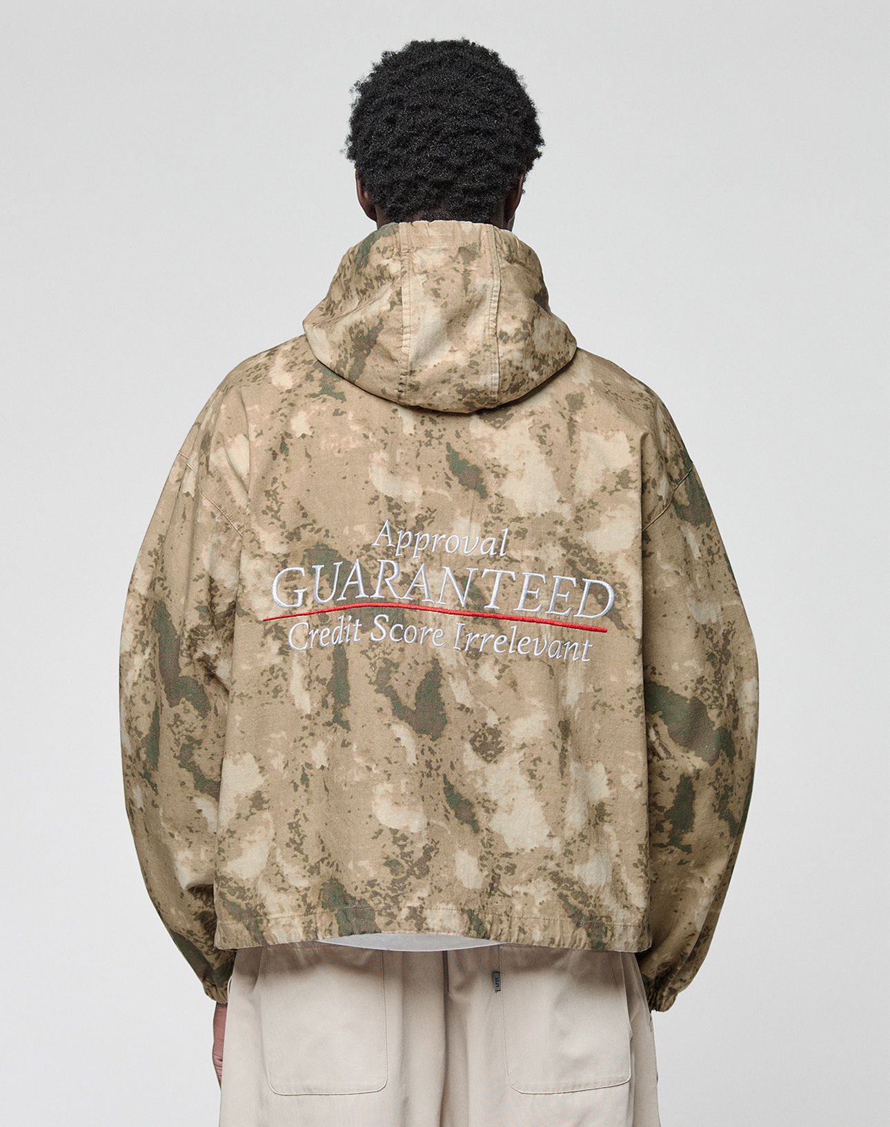 A person is seen from behind wearing the LIVE FAST DIE YOUNG LFDY Camo Halfzip Jacket, an oversize tan and green camouflage piece with a hood and “Approval GUARANTEED Credit Score Irrelevant” on the back, paired with light-colored pants.