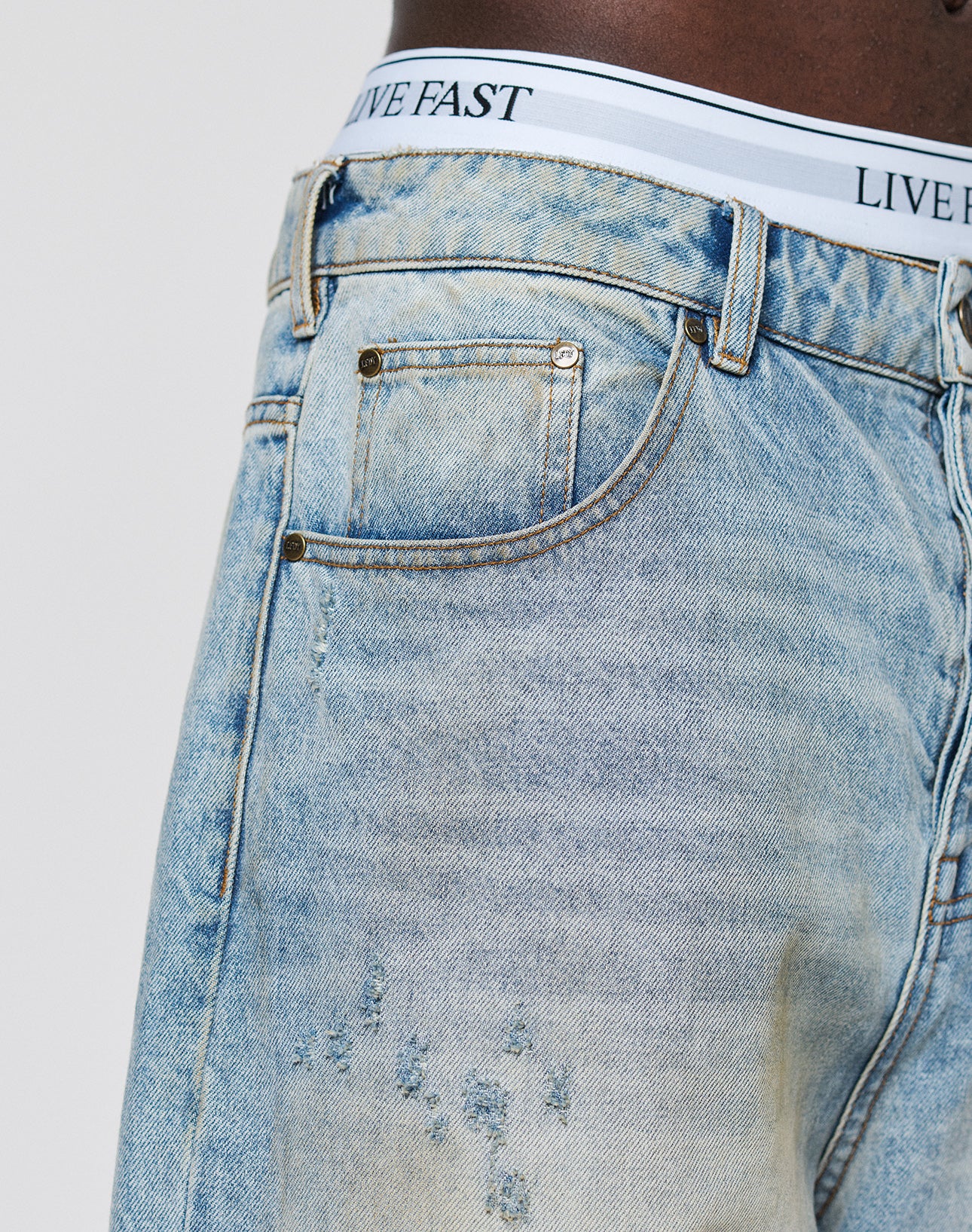 Close-up of a person wearing LFDY Baggy Denim by LIVE FAST DIE YOUNG, styled with white underwear featuring a LIVE FAST waistband. The jeans showcase a relaxed baggy fit, visible pockets, detailed stitching, and thigh distressing.