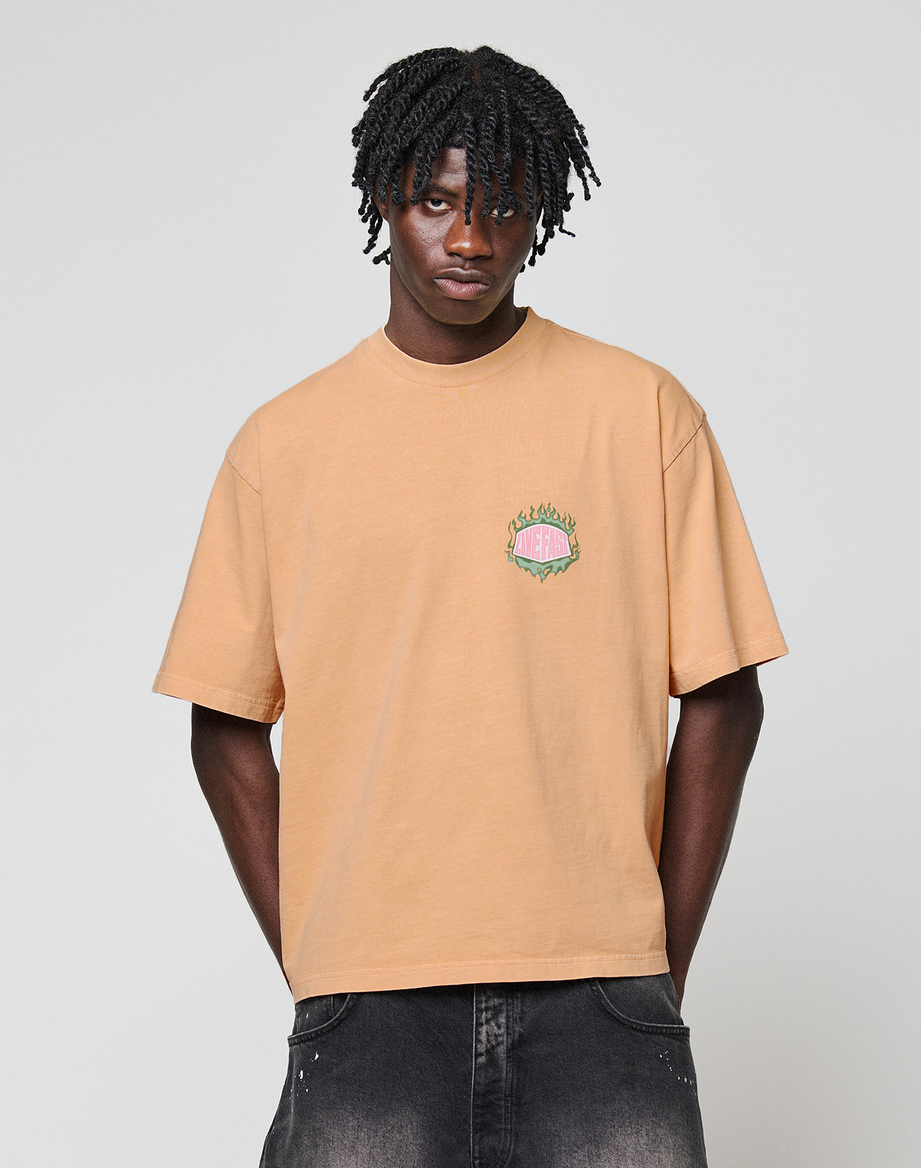 A young man with short twists wears the LFDY Flames Chest Tee from LIVE FAST DIE YOUNG—a loose, light brown 100% Baumwolle shirt with a small, colorful chest frontprint—paired with dark jeans against a plain light gray background.