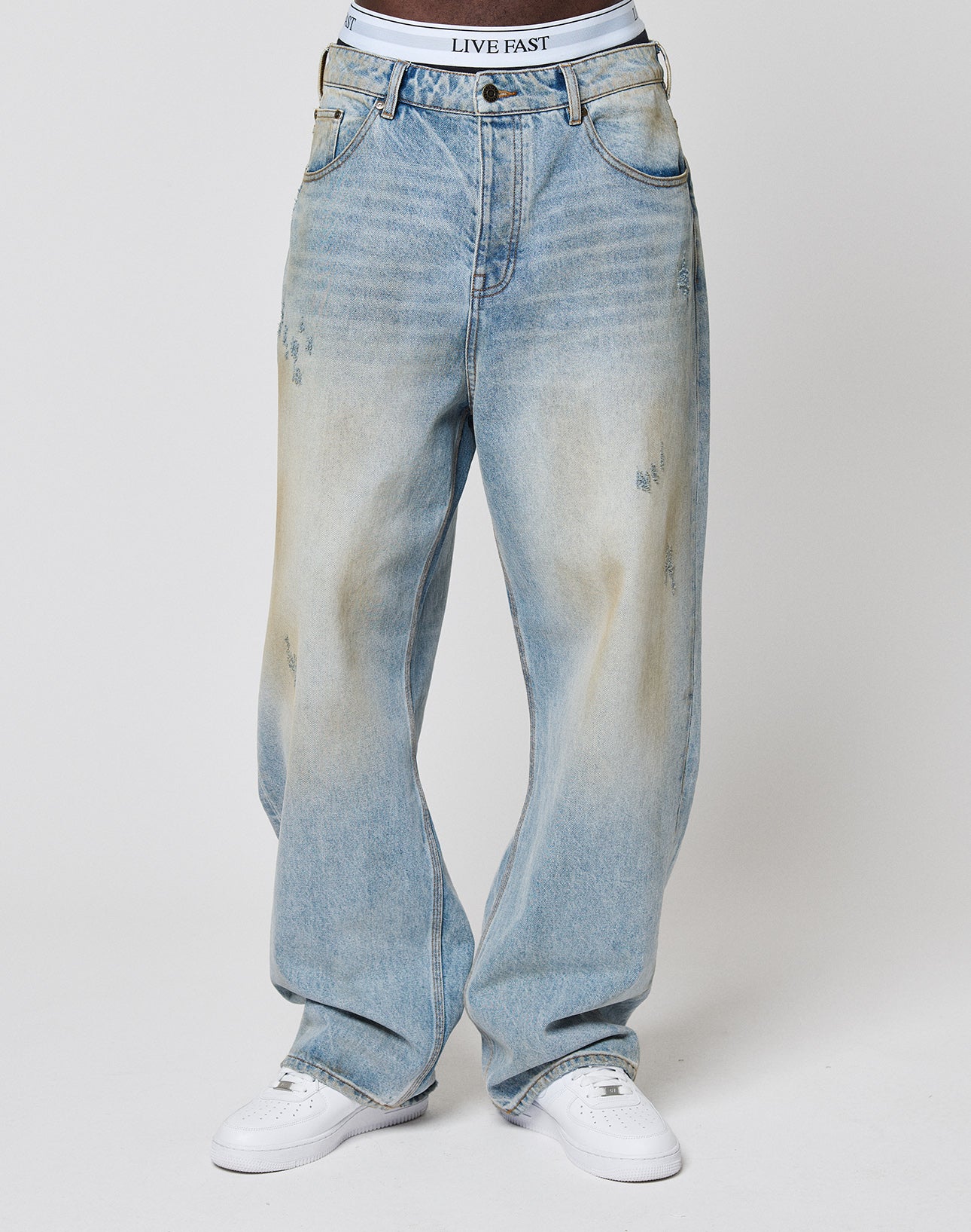A person wears light blue, slightly distressed LFDY Baggy Denim by LIVE FAST DIE YOUNG with white sneakers and a visible LIVE FAST waistband, standing against a plain white background.