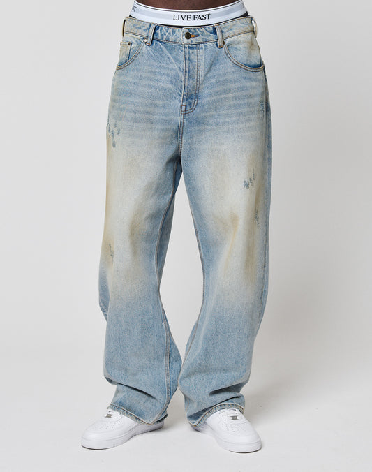 Someone wearing LFDY Baggy Dirt Denim jeans by LIVE FAST DIE YOUNG, styled baggy and faded blue with straight legs, white sneakers, and visible LIVE FAST branded white underwear, stands against a plain background.
