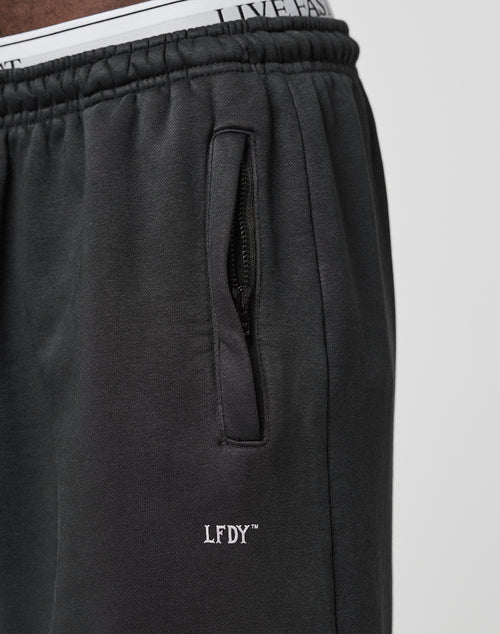 Close-up of a person wearing the Basic Loose Jogger by LIVE FAST DIE YOUNG, featuring a black design, elastic waistband, zippered pocket, white LFDY™ logo print, loose fit, and visible LIVE FAST branded underwear above the waist.