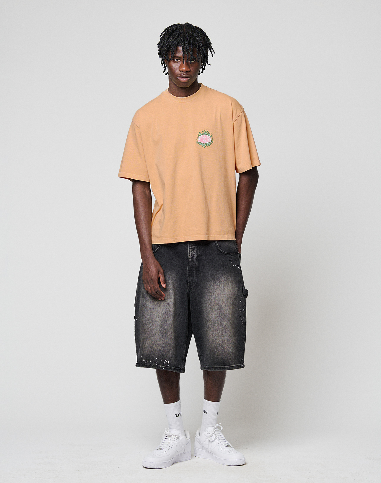 A young man with short dreadlocks stands against a plain background, wearing the LIVE FAST DIE YOUNG LFDY Flames Chest Tee (Regular Fit, 100% Baumwolle), paired with loose black denim shorts, white socks, and white sneakers. His hands are by his sides.