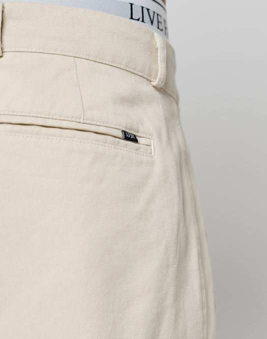 Close-up of the back of beige Wide Chino pants by LIVE FAST DIE YOUNG, in 100% cotton, showing a welt pocket with a black tag, part of a belt loop, and the waistband of LIVE FAST DIE YOUNG underwear visible at the top.